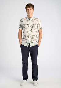 Camisa de botones de manga corta en crema claro con estampado floral en verde y rosa. Combinada con pantalones azul marino oscuro y zapatillas blancas.