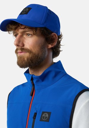 Cappello blu e gilet blu con colletto alto, dotato di una patch nera e una zip rossa. Il tessuto appare liscio e leggero.