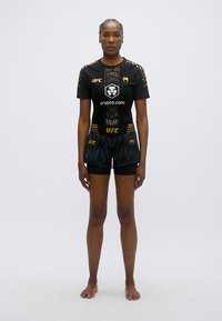 Črna športna obleka z zgornjim delom s kratkimi rokavi in kratkimi hlačami, z zlato obrobo, mrežasto teksturo in UFC blagovno znamko na obeh kosih.