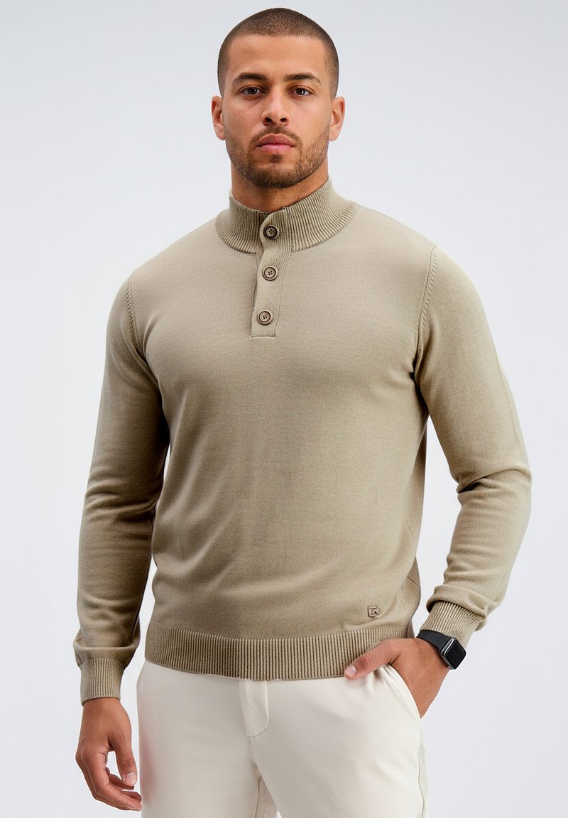 Hombre con suéter de cuello alto beige abotonado y pantalones claros, de pie con una mano en el bolsillo y expresión neutral, contra un fondo liso.