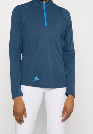 Femme portant un haut de sport bleu marine à manches longues avec une fermeture éclair quart de zip bleu clair et un pantalon blanc, debout devant un fond uni.