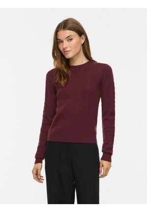 Femme portant un pull cintré à torsades bordeaux à manches longues et un pantalon noir ample sur fond blanc uni.