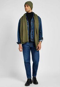 Giacca di jeans, dolcevita nera, jeans blu scuro, sciarpa a coste verde oliva, berretto abbinato e stivali neri con lacci.