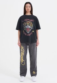 Černé oversized tričko s barevným obrázkem tygra a textem "Los Angeles"; spárované s šedými volnými džíny s žlutým nápisem "Ed Hardy".