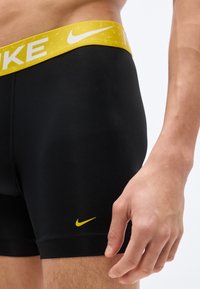 Άνδρας που φοράει μαύρο αθλητικό σορτς με κίτρινη μέση και κίτρινο λογότυπο Nike κοντά στο τελείωμα, στέκεται με χαλαρό το δεξί χέρι.