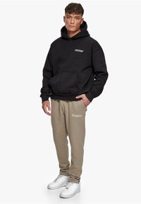 Black hoodie with front pocket, featuring white text. Beige sweatpants with white logo detail. White sneakers complete the outfit.