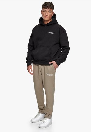 Svart hettegenser med frontlomme, med hvit tekst. Beige joggebukser med hvit logodetalj. Hvite sneakers fullfører antrekket.