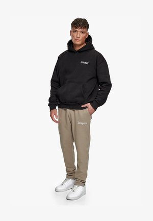 Schwarze Kapuzenjacke mit Fronttasche, die weißen Text enthält. Beigefarbene Jogginghose mit weißem Logodetail. Weiße Sneakers runden das Outfit ab.