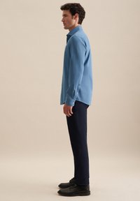 Uomo in piedi di lato, indossa una camicia blu a maniche lunghe con bottoni, pantaloni scuri e scarpe eleganti nere, su uno sfondo semplice.