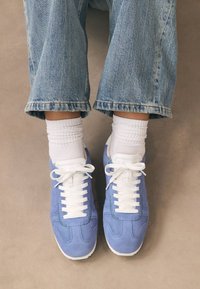 Sneakers in camoscio blu con lacci bianchi e suole di gomma, abbinati a calzini bianchi alla caviglia e jeans leggeri in denim chiaro, larghi e corti.