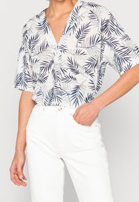 Haut à manches courtes et col en V, en tissu blanc orné de motifs de feuilles bleues. Associé à un jean blanc taille haute. Coupe décontractée et design texturé.