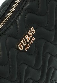 Borsa nera in ecopelle trapuntata con logo dorato "GUESS EST 1981". Presenta una cerniera liscia, un motivo ondulato distintivo e una finitura lucida.