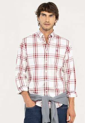 Hombre que lleva una camisa de cuadros blanca y roja con las mangas remangadas, un jersey gris atado a la cintura y vaqueros azules.