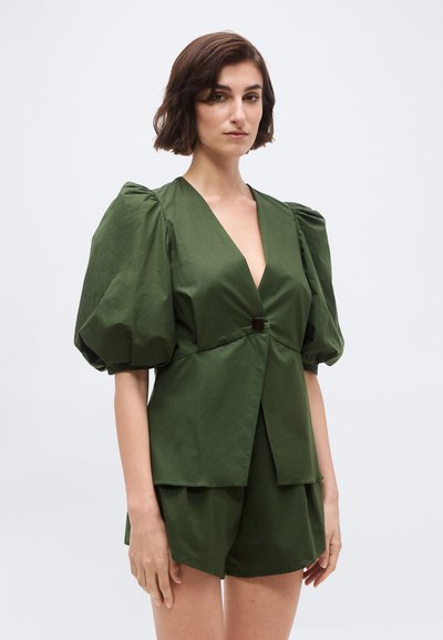 Γυναίκα με κοντά καστανά μαλλιά, που φοράει φόρεμα ελιάς με βαθύ V και φουσκωτά μανίκια, καθώς και σορτς σε ίδια απόχρωση, στέκεται μπροστά σε απλό φόντο.