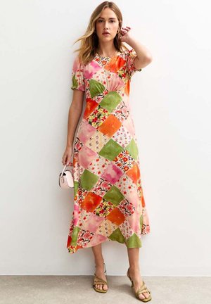 Femme portant une robe midi en patchwork colorée avec des motifs floraux et géométriques, tenant un petit sac à main blanc et portant des talons verts.