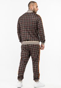 Plaid tracksuit in dark brown, orange accent stripes, elastic cuffs, and waistband. Styled with white sneakers, showcasing a relaxed fit.