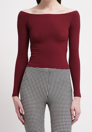 Femme portant un haut bordeaux ajusté à côtes et épaules dénudées, ainsi qu'un pantalon taille haute en pied-de-poule noir et blanc, sur un fond uni.