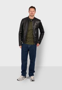 Chaqueta de cuero negra con un cuello redondo y detalles de cremallera, combinada con una camiseta de color verde oliva y vaqueros de mezclilla azul. Unas zapatillas completan el look.