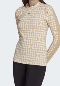 Světle béžový dlouhý rukáv top s kostkovaným vzorem a jemnými květinovými akcenty, s vysokým límcem a přiléhavým střihem. Zlatý detail loga.