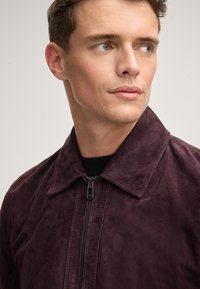 Giacca di suede borgogna con colletto, chiusura zip frontale e dettagli in maglia nera a coste attorno al collo. Texture liscia e vestibilità su misura.