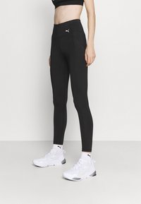 Svarta atletiska leggings med hög midja, med platta sömmar och en Puma-logotyp. Kombinerade med vita sneakers och accentuerade med atletiska strumpor.