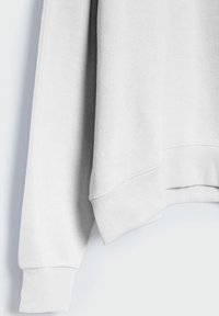 Gros plan sur la manche et l’ourlet d’un sweat-shirt blanc avec des poignets côtelés et des coutures visibles sur un fond clair.