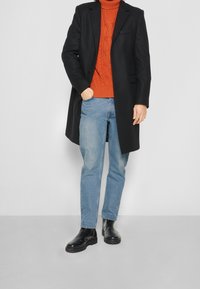Abrigo de lana negro sobre un suéter de cuello alto de punto naranja, combinado con pantalones vaqueros de denim azul claro y botas de cuero negras por el tobillo. Texturas suaves y corte entallado.