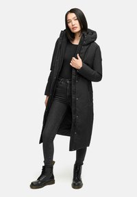 Svart lång parka med huva, framknappar och sidofickor. Bärs över en svart topp och slim fit-jeans, ihop med svarta snörstövlar.