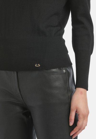 Czarny dzianinowy sweter z prążkowanym wykończeniem na dole i długimi rękawami, w zestawieniu z czarnymi skórzanymi spodniami. Na dole swetra znajduje się małe złote logo.