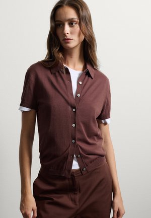 Cardigan corto marrone a maniche corte con chiusura a bottoni, colletto e polsini a costine, indossato sopra una camicia bianca. Tessuto morbido, design casual.