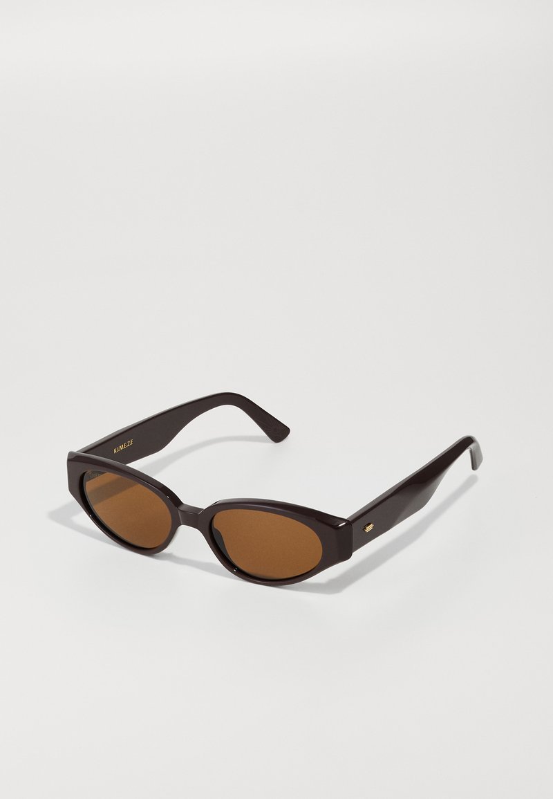 Faux cat-eye solbriller med mørkebrune plastikrammer og brune linser. Har guld logo detaljer på armen og en glat, blank overflade.
