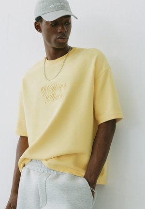 Junger Mann trägt ein gelbes Shirt mit dem Text "Walking Places", graue Jogginghose, graue Kappe und silberne Kette, steht mit den Händen in den Taschen.