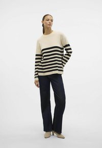 Maglione lavorato a maglia di colore crema con righe orizzontali nere, taglio rilassato, scollo rotondo. Abbinato a jeans a gamba larga blu scuro e scarpe con tacco beige.