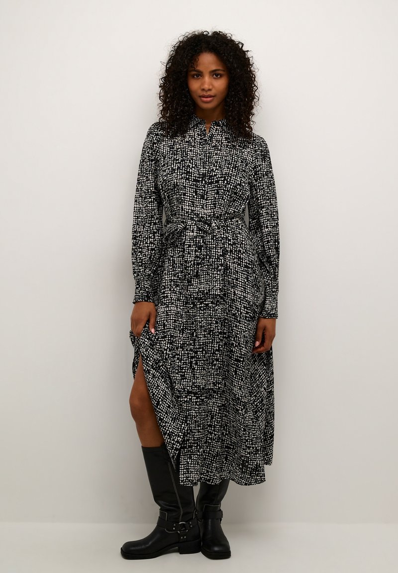 Robe à motif noir et blanc avec des manches longues, fermeture boutonnée à l'avant et ceinture à la taille ; présente des fentes sur les côtés et une silhouette fluide.