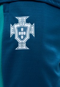 Μπλε αθλητικό ύφασμα με λευκό έμβλημα που απεικονίζει μια ασπίδα με πέντε μικρότερες ασπίδες και τα γράμματα F.P.F., στο κέντρο ενός σχήματος σταυρού.