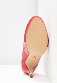 Chaussure à talons hauts rouge vue de la semelle avec une semelle beige lisse et un talon fin, positionnée devant un fond d'angle blanc.