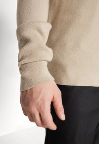 Beige kötött pulóver bordázott mandzsettával és sima textúrával, fekete nadrággal párosítva. A kéz a mandzsetta közelében helyezkedik el, kiemelve a részleteket.