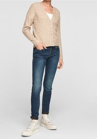Beige žebrovaný cardigan s výstřihem do V a třemi knoflíky, v kombinaci s tmavě modrými skinny džíny a bílými platformovými teniskami.