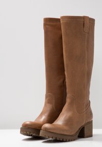 Botas altas hasta la rodilla de cuero marrón con acabado texturizado, tacón cuadrado y suela de goma. Presentan un diseño cosido y una lengüeta para tirar en la parte superior.