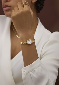 Orologio da polso oro con quadrante rotondo, quadrante bianco, numeri argentati e bracciale a maglie. Indossato su un polso con una blusa bianca.