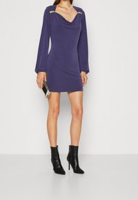 Fialové šaty s rolákem a dlouhými rukávy, zdobené zlatými knoflíky na ramenou; strukturovaná tkanina a přiléhavý střih; doplněné černými kotníkovými botami.