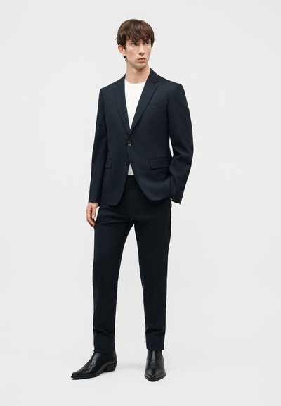 Νεαρός άνδρας όρθιος, φοράει μαύρο σακάκι και παντελόνι, λευκό πουκάμισο και μαύρα δερμάτινα μποτάκια, σε φωτεινό απλό φόντο.