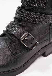 Botín negro de cuero hasta el tobillo con superficie texturizada, hebilla de metal y correas con tachuelas. Cuenta con un tacón grueso y un diseño de cordones en el frente.