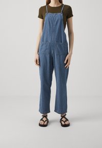 Nainen, joka on pukeutunut siniraitaisiin ja valkoisiin pystysuoriin farkkuhousuihin, oliivinvihreään lyhythihaiseen paitaan ja mustiin hihnakenkiin, seisoo valkoista taustaa vasten.