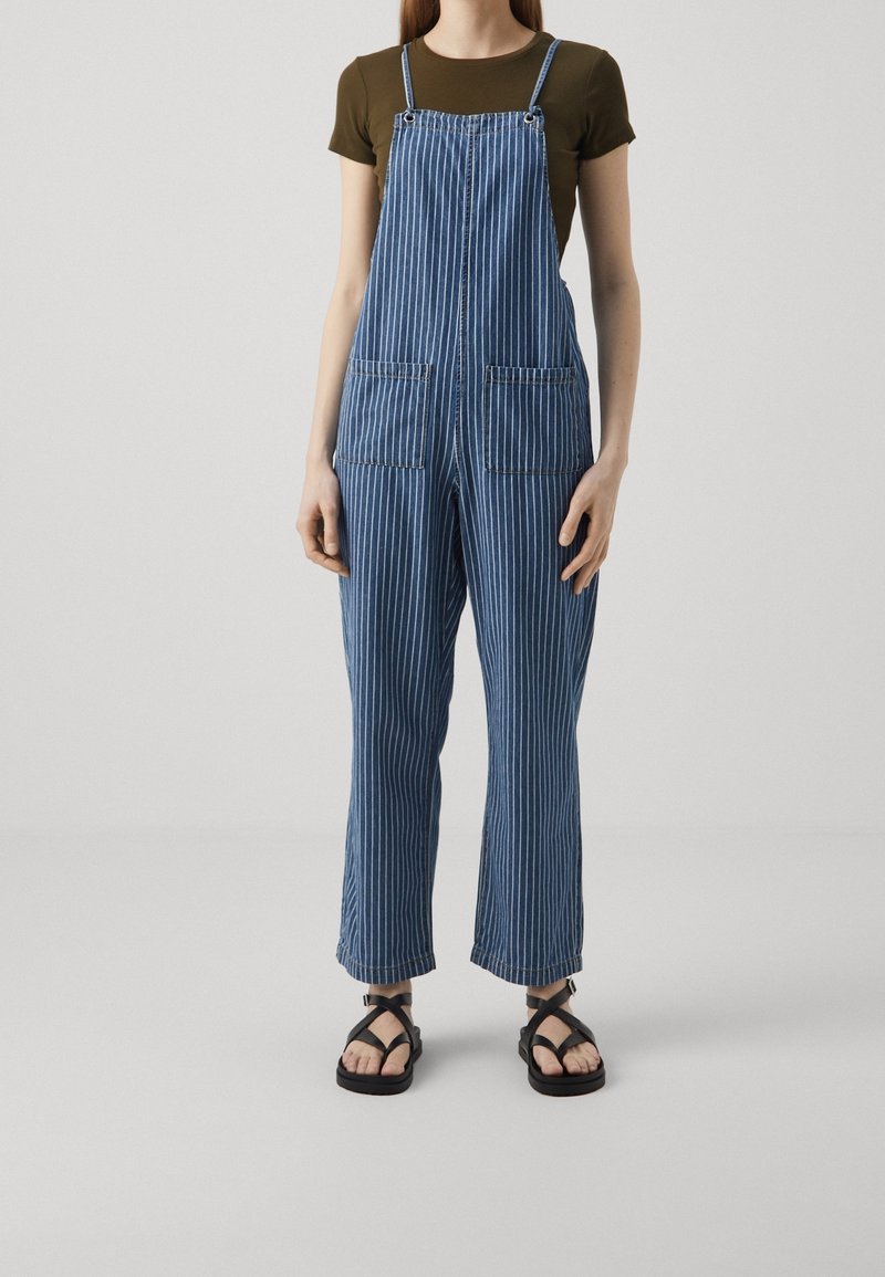 Nainen, joka on pukeutunut siniraitaisiin ja valkoisiin pystysuoriin farkkuhousuihin, oliivinvihreään lyhythihaiseen paitaan ja mustiin hihnakenkiin, seisoo valkoista taustaa vasten.