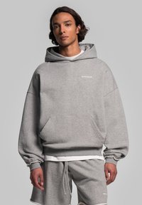 Felpa grigia oversize realizzata in tessuto morbido, con tasca frontale e cappuccio con lacci, caratterizzata da dettagli del logo bianco sul petto.