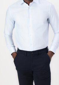 Světle modrá košile na knoflíky s dlouhými rukávy, spárovaná s tmavě modrými kalhotami. Košile má jemnou texturu a standardní design límce.