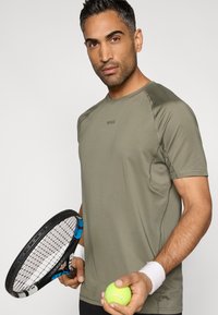 Oliwkowo-zielona sportowa koszulka z logo, krótkie rękawy, bezszwowy design, w zestawieniu z rakietą tenisową i neonowo żółtą piłką.