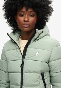 Giacca a piumino verde chiaro con chiusura a zip, dettagli neri, cappuccio e logo sul petto, caratterizzata da sezioni trapuntate e interno morbido.