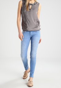 Vrouw in een mouwloos grijs top met een bewerkte halslijn, lichtblauwe skinny jeans en beige tassel loafers, staand tegen een witte achtergrond.